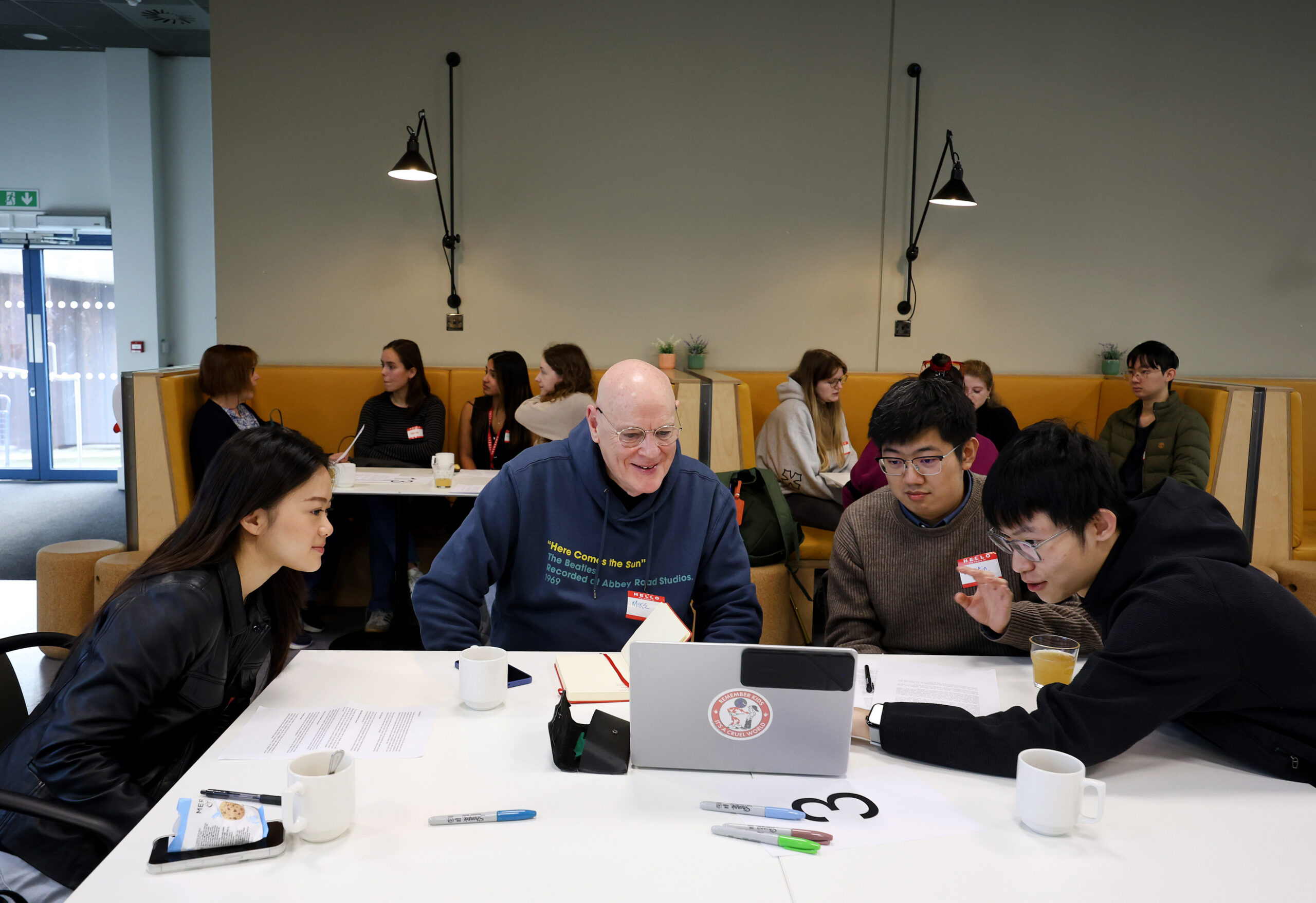 260130 Science Communication workshop 2475 'Three diverse student and a patient are sitting at a table, gathered around a laptop. Another student is pointing at the screen, explaining something. All the students are smiling. Strewn on the table are items such as cups, coloured felt pens and notes. Behind them are two booths with other seating and each booth has a table. Diverse students and a patient are sat at each table, talking and smiling. The tables are strewn with similar items