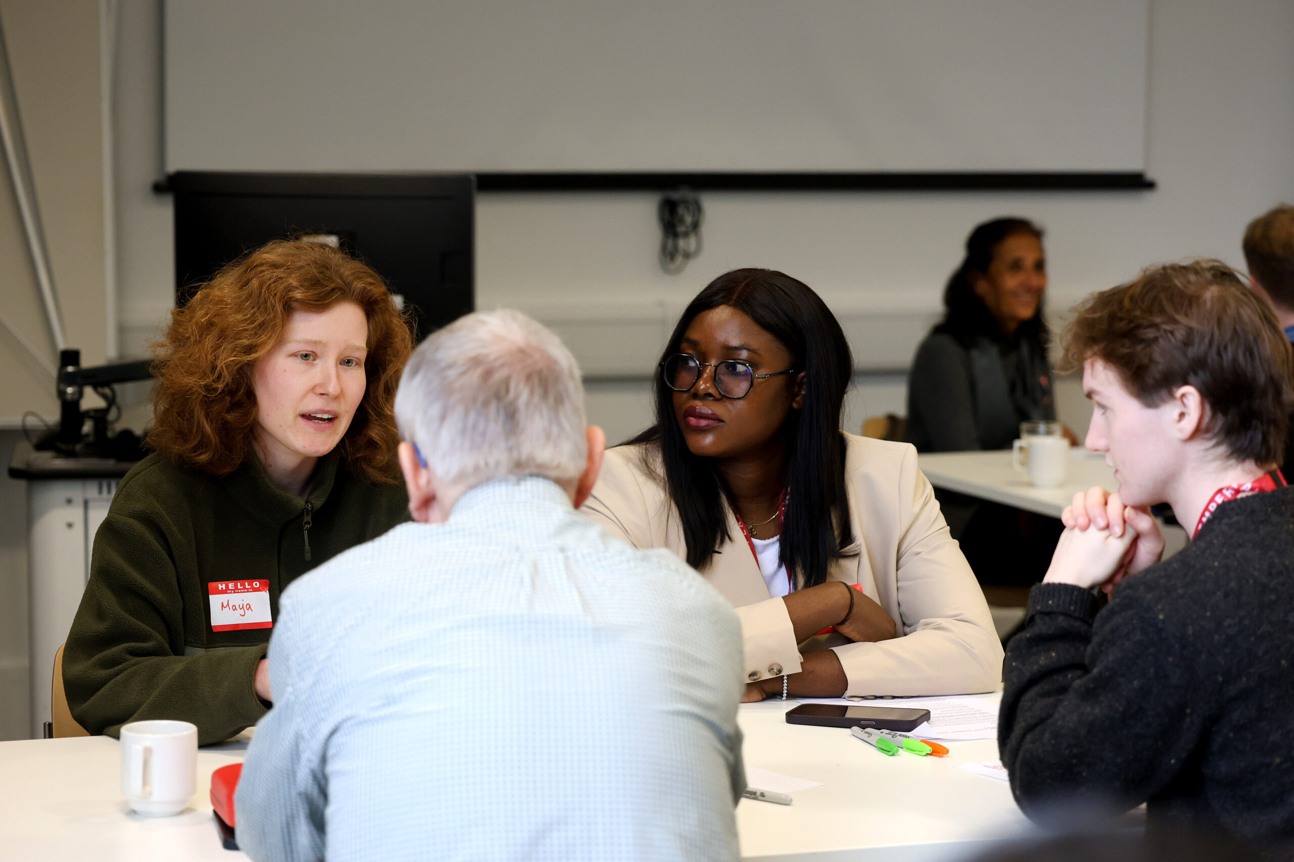 260130 Science Communication workshop 2603 'Three diverse students and a patient, sitting around a table, deep in conversation. There are pens, notes, cups and phones scattered on the table. The background is largely blurred out but you can make out a projector, a laptop on a stand and another group sitting at a separate table